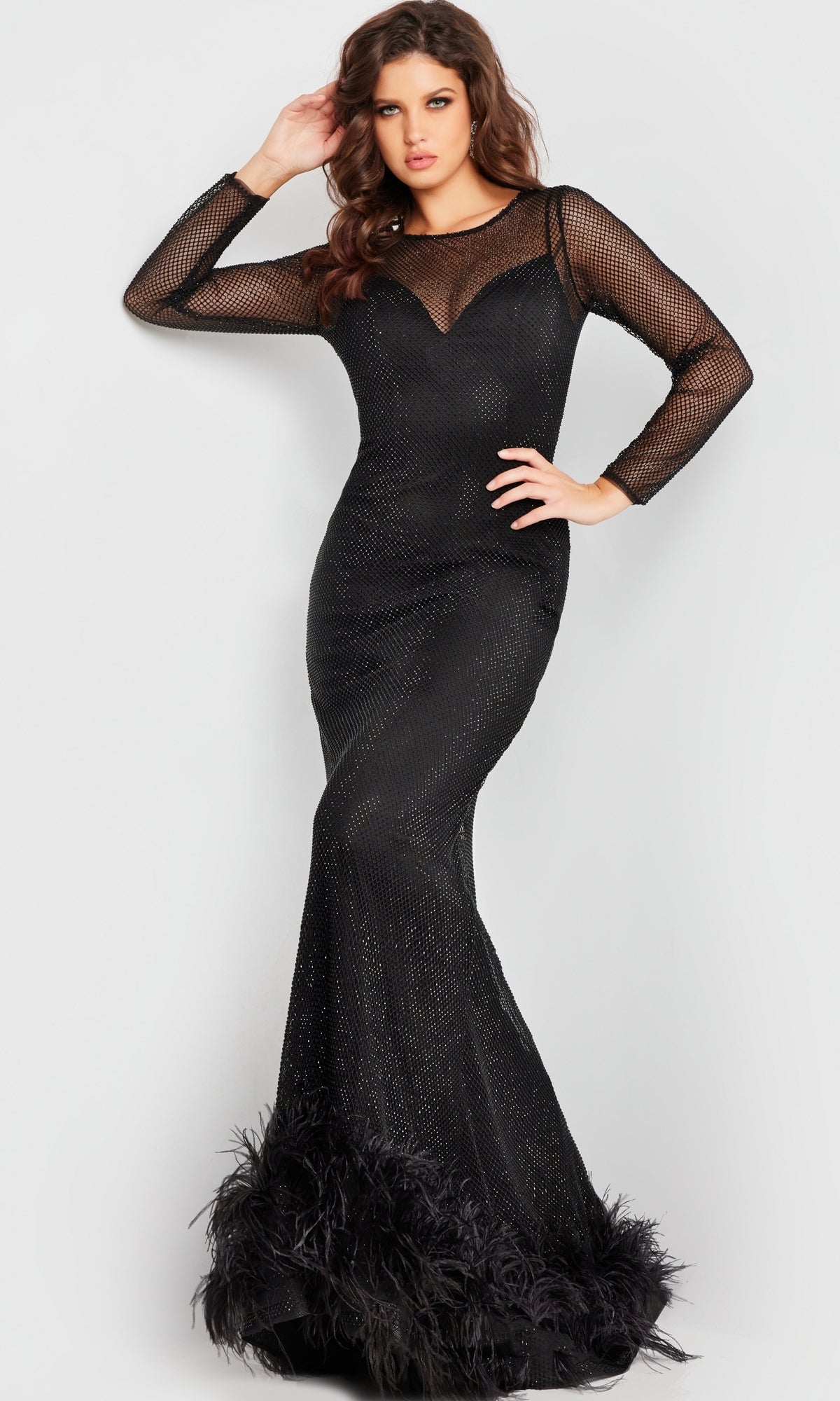 A woman stands against a plain light background wearing the Jovani Long Sleeve Long Black Formal Dress 26047, featuring sheer mesh sleeves, subtle glitter details, and feathered trim at the hem.